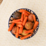 Overhead view of a blue and white patterned bowl filled with roasted baby carrots and small red and purple potatoes, seasoned with visible spices, set on a cream textured cloth atop a wood table.
