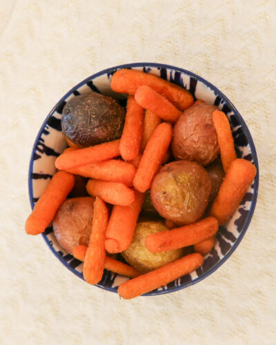 Overhead view of a blue and white patterned bowl filled with roasted baby carrots and small red and purple potatoes, seasoned with visible spices, set on a cream textured cloth atop a wood table.