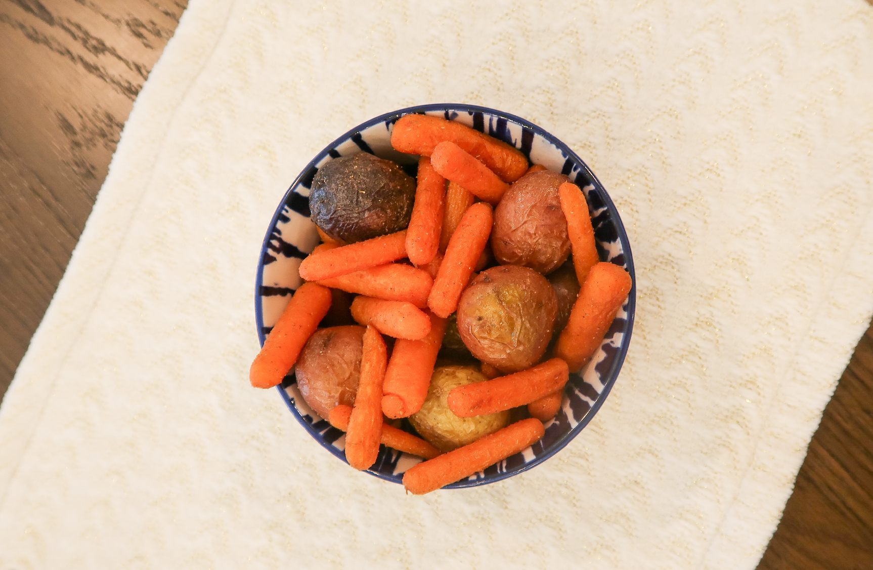 Overhead view of a blue and white patterned bowl filled with roasted baby carrots and small red and purple potatoes, seasoned with visible spices, set on a cream textured cloth atop a wood table.
