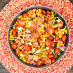 Overhead view of a metal bowl filled with a corn and bean salad mixed with halved cherry tomatoes, diced zucchini, and red onion, placed on a red and white woven placemat on a wood table.
