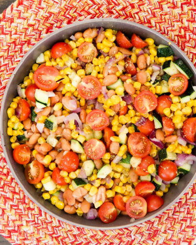Overhead view of a metal bowl filled with a corn and bean salad mixed with halved cherry tomatoes, diced zucchini, and red onion, placed on a red and white woven placemat on a wood table.