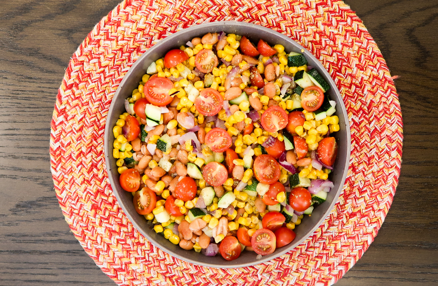 Overhead view of a metal bowl filled with a corn and bean salad mixed with halved cherry tomatoes, diced zucchini, and red onion, placed on a red and white woven placemat on a wood table.