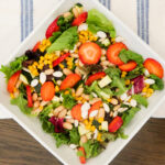 Overhead view of a square white bowl containing a mixed greens salad topped with sliced strawberries, corn, white beans, diced zucchini, and sliced almonds, set on a blue and white striped cloth.