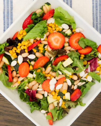 Overhead view of a square white bowl containing a mixed greens salad topped with sliced strawberries, corn, white beans, diced zucchini, and sliced almonds, set on a blue and white striped cloth.