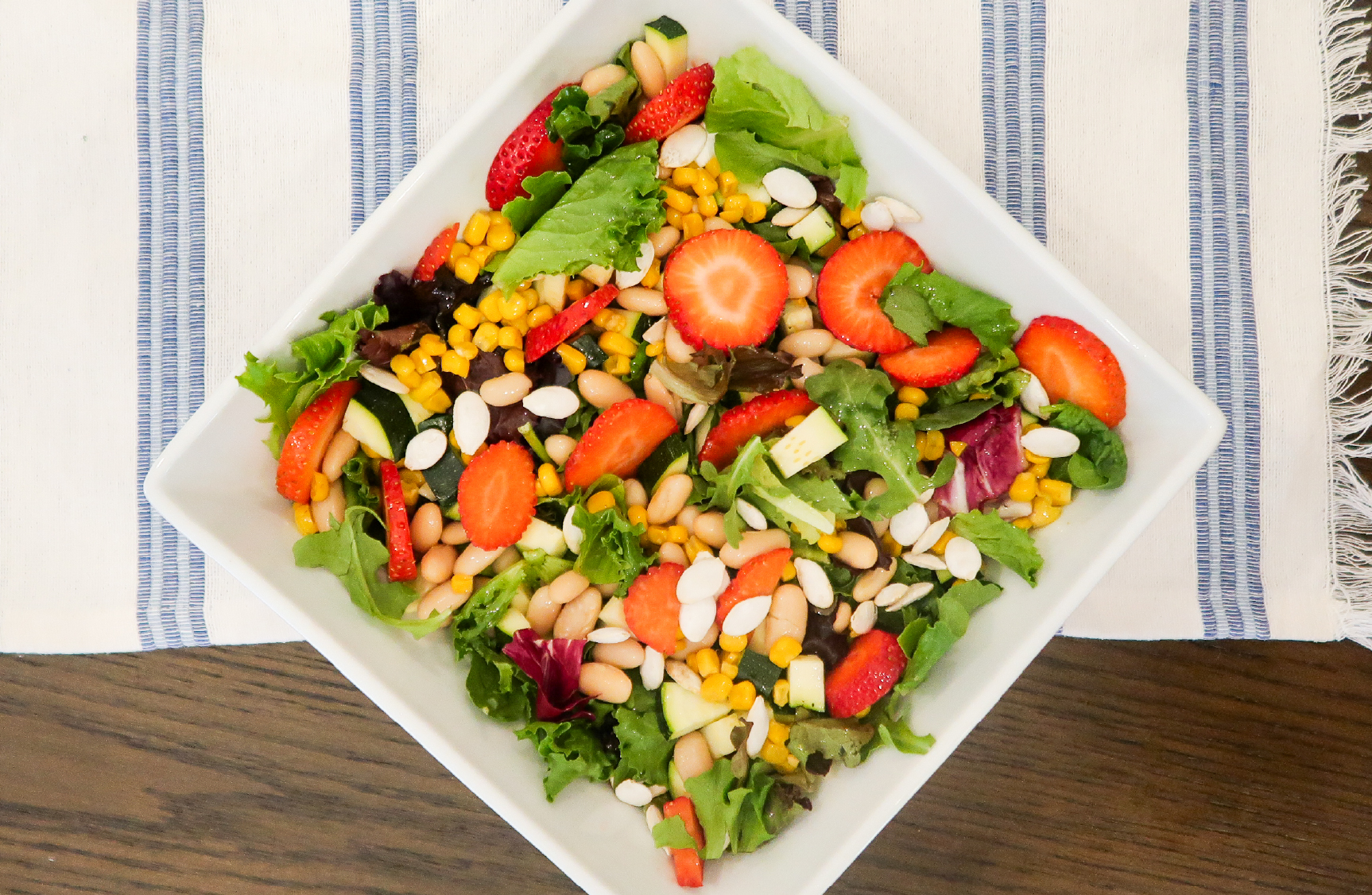 Overhead view of a square white bowl containing a mixed greens salad topped with sliced strawberries, corn, white beans, diced zucchini, and sliced almonds, set on a blue and white striped cloth.
